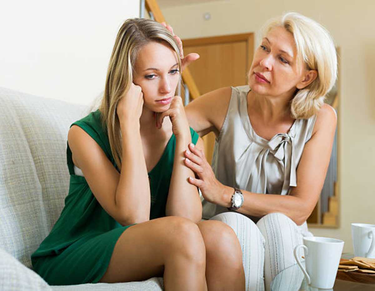Two women sit on a couch at home; a younger woman looks upset with her head resting on her hands while an older woman gently touches her arm and appears to comfort her.