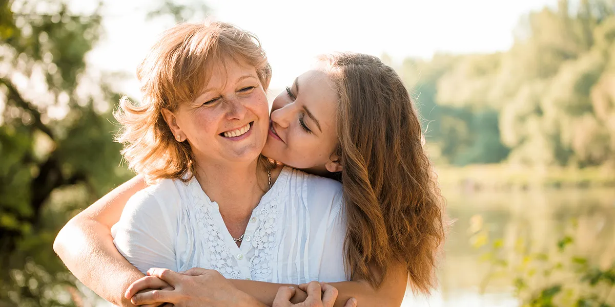 A smiling older woman is hugged from behind by a younger woman, who kisses her cheek, as they enjoy a sunny moment outdoors beside a lake.