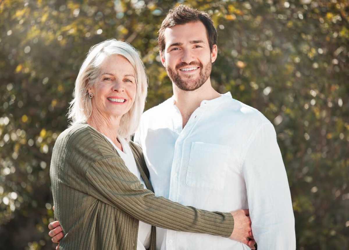An older woman with gray hair smiles and embraces a younger bearded man in a white shirt as they pose together outdoors in warm sunlight.