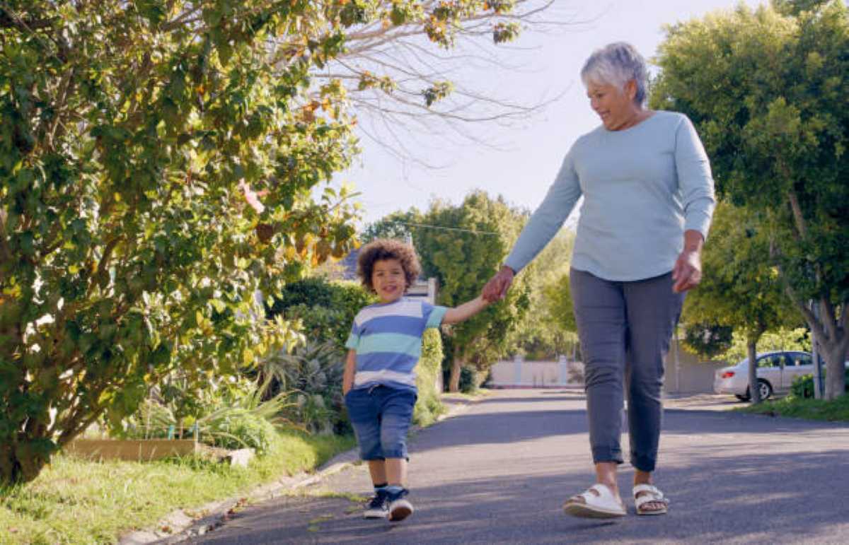 A smiling grandmother holds hands with her young grandson as they walk together down a quiet, tree-lined neighborhood street on a sunny day.