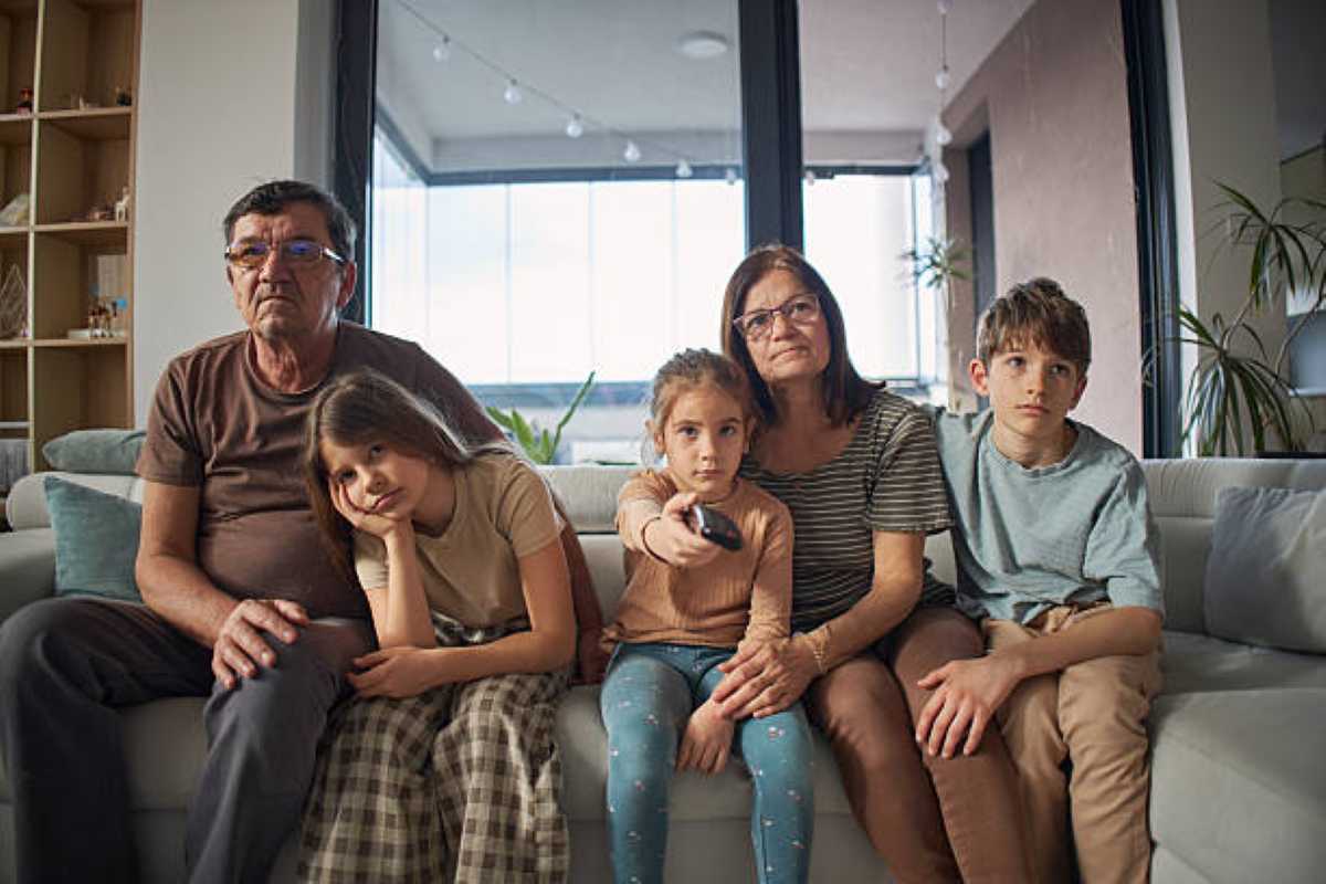 Grandparents and three grandchildren sitting together on a living room couch, looking bored and disconnected during a family visit.