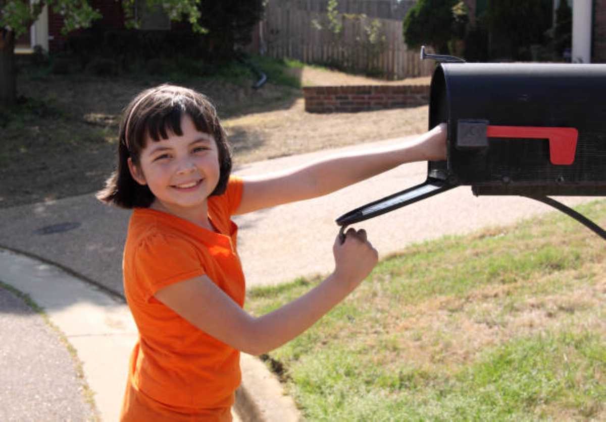 Smiling young girl in an orange shirt opening a black mailbox on a sunny day, excited to check for a letter or special surprise in the mail.
