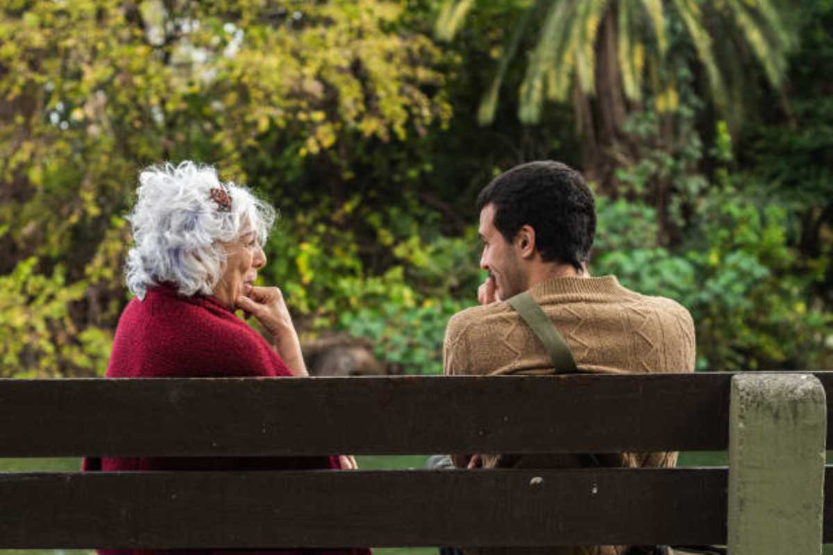 An elderly grandmother with curly white hair and a red sweater sits on a wooden park bench, having a heartfelt conversation with her adult grandson in a peaceful, green park.