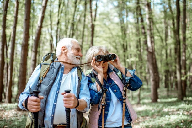 7 Easy Hobbies the Happiest Women Over 60 Are Falling in Love With 8 A senior couple enjoys a nature walk through a lush green forest. The man carries a backpack and trekking poles while the woman looks through binoculars, observing wildlife.