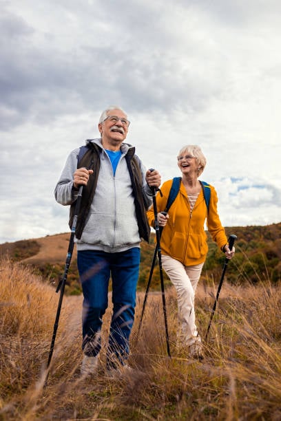 7 Easy Hobbies the Happiest Women Over 60 Are Falling in Love With 5 A cheerful older couple hikes together across a grassy hillside under a cloudy sky, smiling as they walk with trekking poles and backpacks.