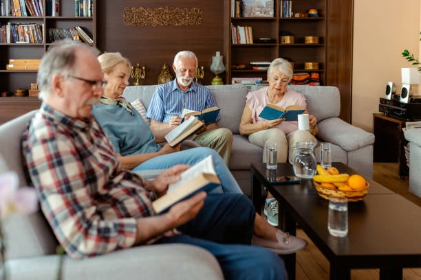 7 Easy Hobbies the Happiest Women Over 60 Are Falling in Love With 9 Four older adults sit comfortably in a cozy living room, quietly reading books together, surrounded by bookshelves, warm furnishings, and refreshments.