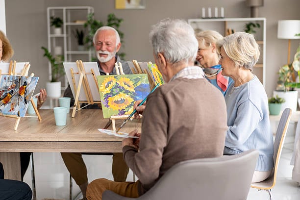 7 Easy Hobbies the Happiest Women Over 60 Are Falling in Love With 3 A group of senior adults enjoys an indoor painting class, creating colorful artwork on small easels while chatting and sharing a relaxing creative experience.