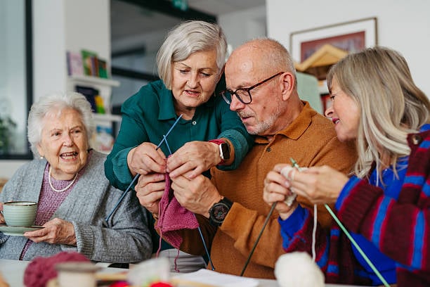 7 Easy Hobbies the Happiest Women Over 60 Are Falling in Love With 7 Four older adults gather around a table for a knitting session. One woman helps a man with his knitting project while the others smile and work with colorful yarn, enjoying a friendly social activity.