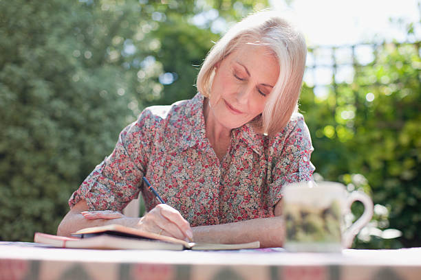 7 Easy Hobbies the Happiest Women Over 60 Are Falling in Love With 6 An older woman with short silver hair and a floral blouse smiles gently while writing in a notebook at an outdoor table, with a ceramic mug beside her and a lush green garden in the background.