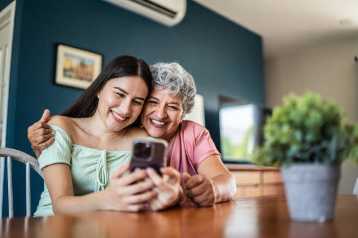 A smiling grandmother with short gray hair and a pink shirt sits close to her granddaughter, who wears a light green top, as they happily look at a smartphone together at a wooden table in a cozy, sunlit home.