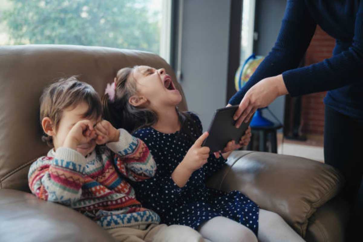 Two young children sitting on a couch, one crying and rubbing his eyes while the other looks upset and holds a tablet as an adult reaches to take it away.