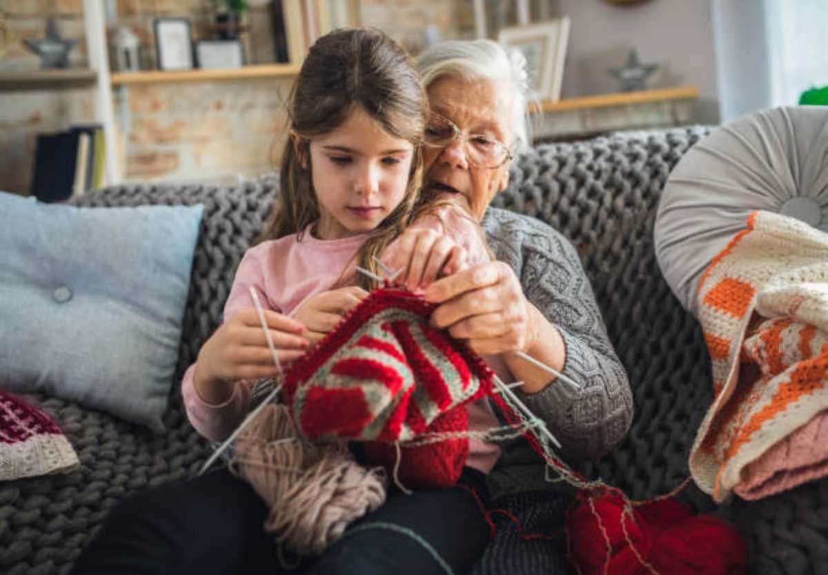 Alt text A grandmother teaches her young granddaughter how to knit a colorful red and white project as they sit together on a cozy couch, sharing a special bonding moment at home.
