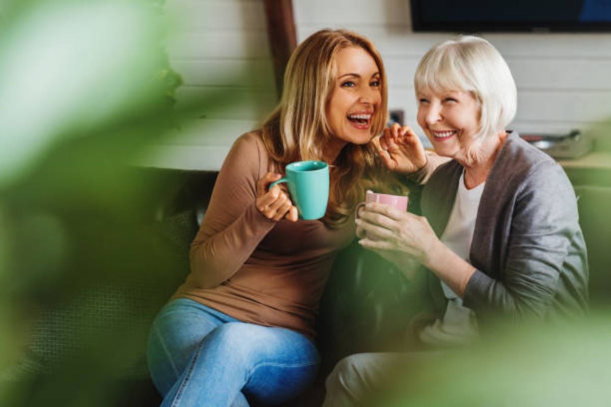 A smiling elderly mother and her adult daughter share a joyful moment over coffee on the couch, highlighting the importance of love, communication, and staying connected with grown children.