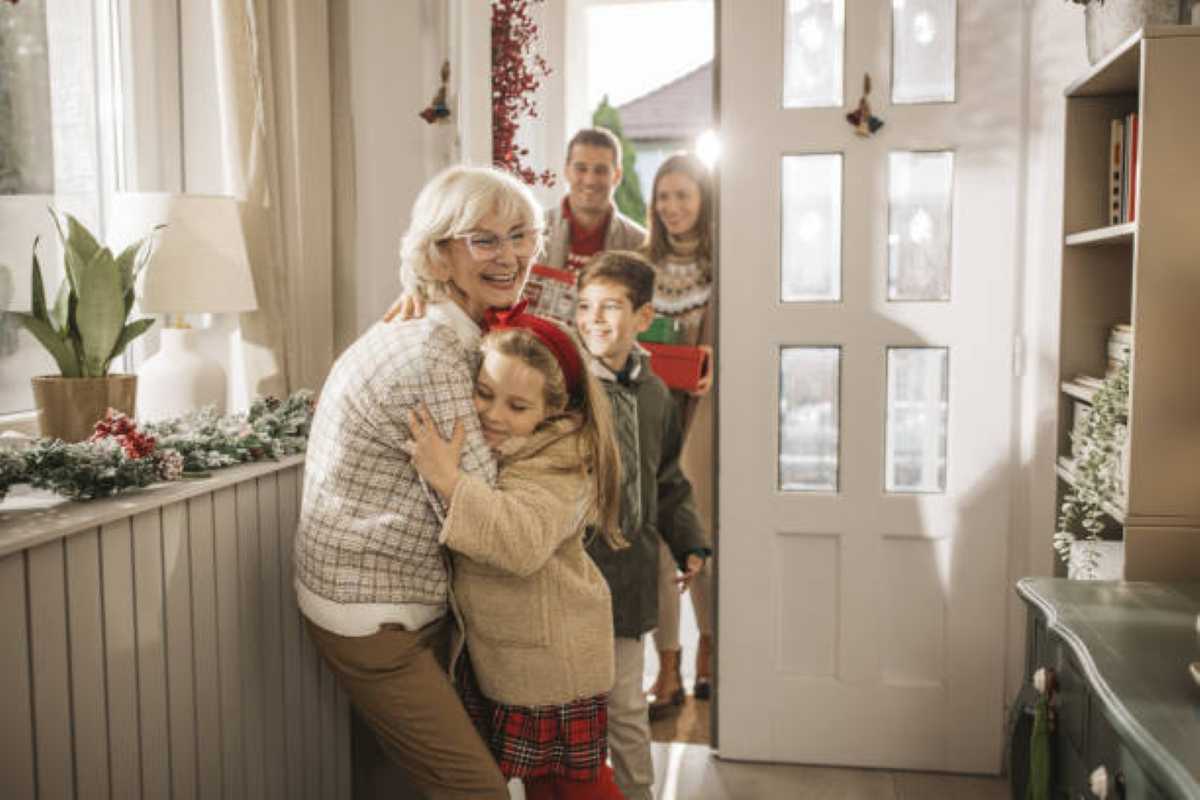 Grandmother hugging her young granddaughter at the front door while other family members arrive behind them carrying gifts.