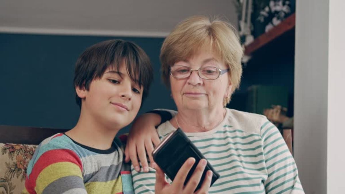 A grandmother and her young grandson sit close together on a couch, looking at a smartphone and enjoying a quiet moment of connection at home.