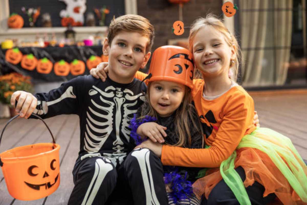Three smiling children dressed in colorful Halloween costumes pose together outdoors. One wears a black skeleton outfit and holds an orange pumpkin candy bucket, the youngest wears an orange pumpkin bucket as a hat, and the third wears a pumpkin-themed costume with a bright green and orange tutu. Halloween decorations hang in the background.