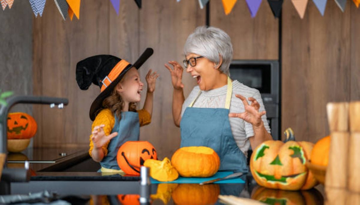 A smiling grandmother with short gray hair and glasses and her young granddaughter wearing a black-and-orange witch hat playfully raise their hands while decorating pumpkins together in a cozy kitchen. Both wear blue aprons and appear excited, surrounded by carved jack-o'-lanterns, colorful pumpkins, and festive Halloween bunting.