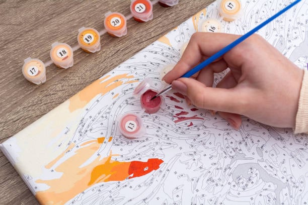 Remember Paint-by-Numbers From the 1960s? You Won’t Believe What They Look Like Today 4 A close-up of a hand using a thin blue paintbrush to fill numbered sections of a paint-by-numbers canvas with red paint. Small numbered paint pots and partially painted orange and yellow areas are visible on a wooden table.