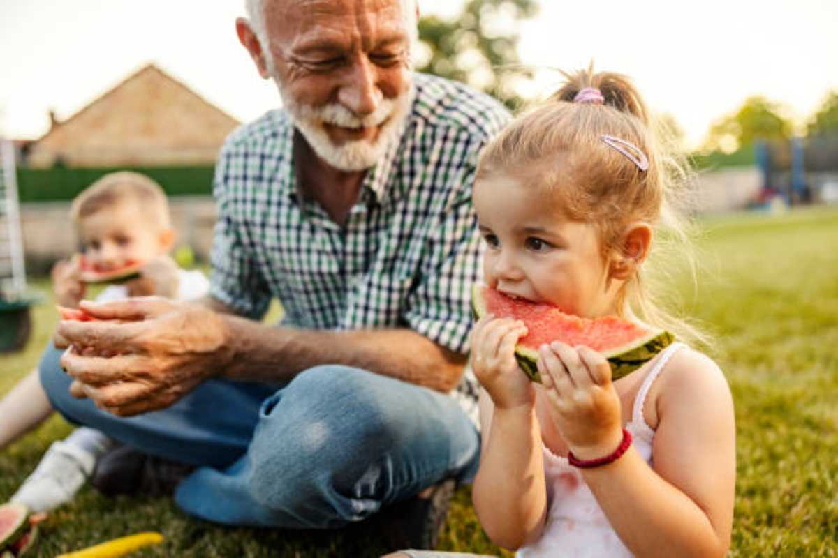 An elderly grandfather sits on the grass outdoors with two young grandchildren, smiling as they enjoy slices of watermelon together. In the foreground, a little girl holds a large slice of watermelon, while a younger child eats in the background. The scene captures a warm family moment on a sunny day.