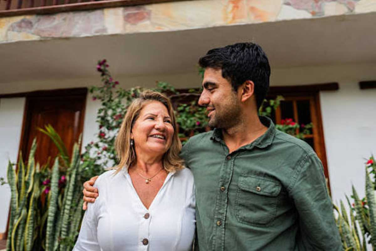 A smiling mother and her adult son stand close together with their arms around each other outside a home.