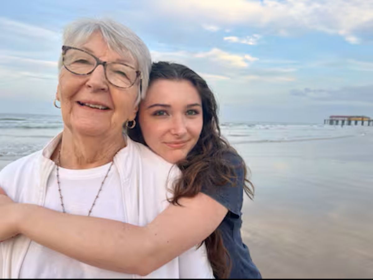 A smiling grandmother with short white hair and glasses shares a warm hug with her teenage granddaughter on a peaceful beach, with ocean waves and a soft evening sky in the background.