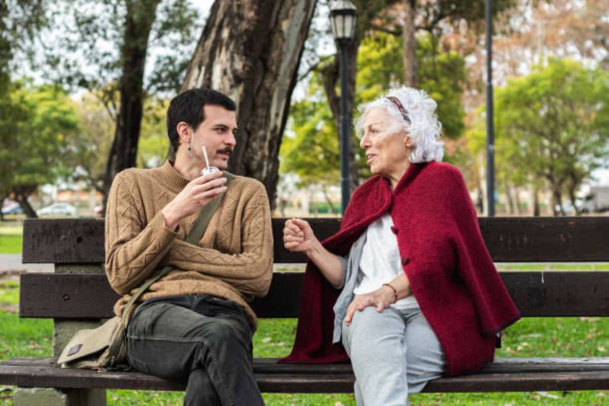 A grandmother in a red shawl shares a warm conversation with her adult grandson as they sit together on a wooden park bench surrounded by trees and greenery.