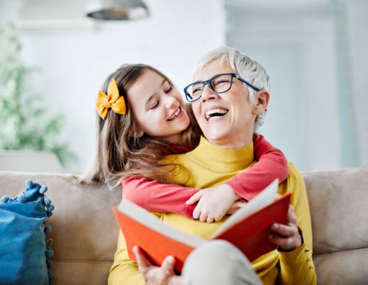 Give me the alt text A smiling grandmother with short gray hair and glasses reads a book while her granddaughter hugs her from behind on the couch.
