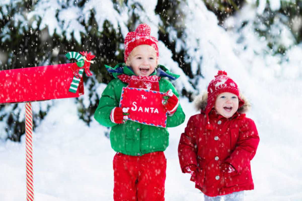Two smiling young children dressed in festive red and green winter outfits stand in falling snow beside a red mailbox, with one holding a letter to Santa in front of snow-covered evergreen trees.