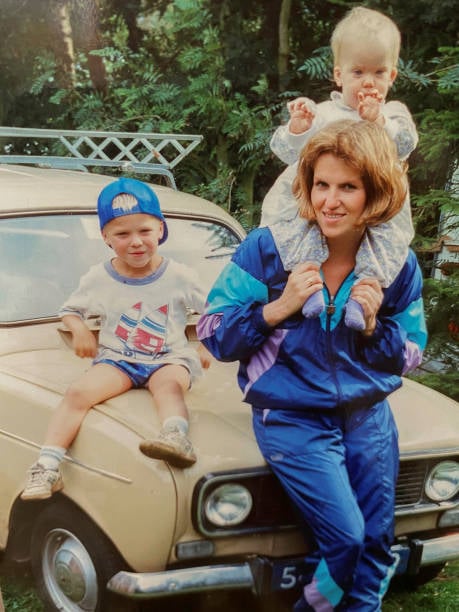 15 Unexpected Things to Mail Your Grandkids That’ll Make Them Smile From Ear to Ear 4 Grandmother posing by a vintage car with one young grandson beside her and a baby grandchild on her shoulders.