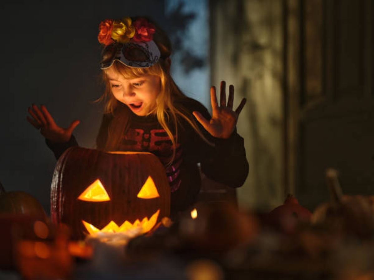 A young girl wearing a colorful floral headband and a skeleton-themed Halloween outfit reacts with excitement to a glowing jack-o'-lantern. Her mouth is open in surprise, and her hands are raised as warm orange light from the carved pumpkin illuminates her face. Several other pumpkins surround her in the dimly lit, festive setting.