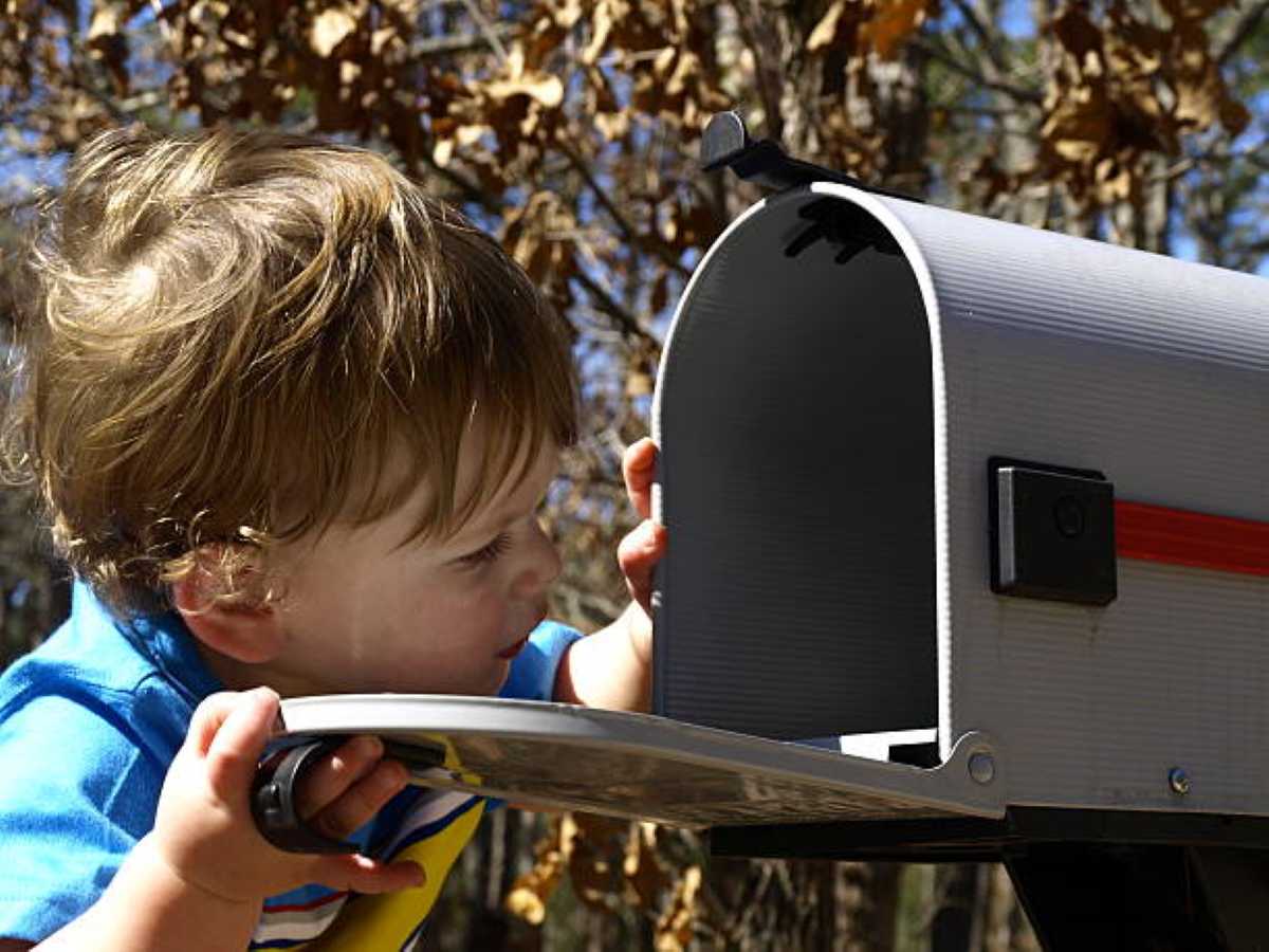 A young boy in a bright blue shirt eagerly peeks inside an open mailbox on a sunny day, excited to see if a special letter or surprise has arrived.